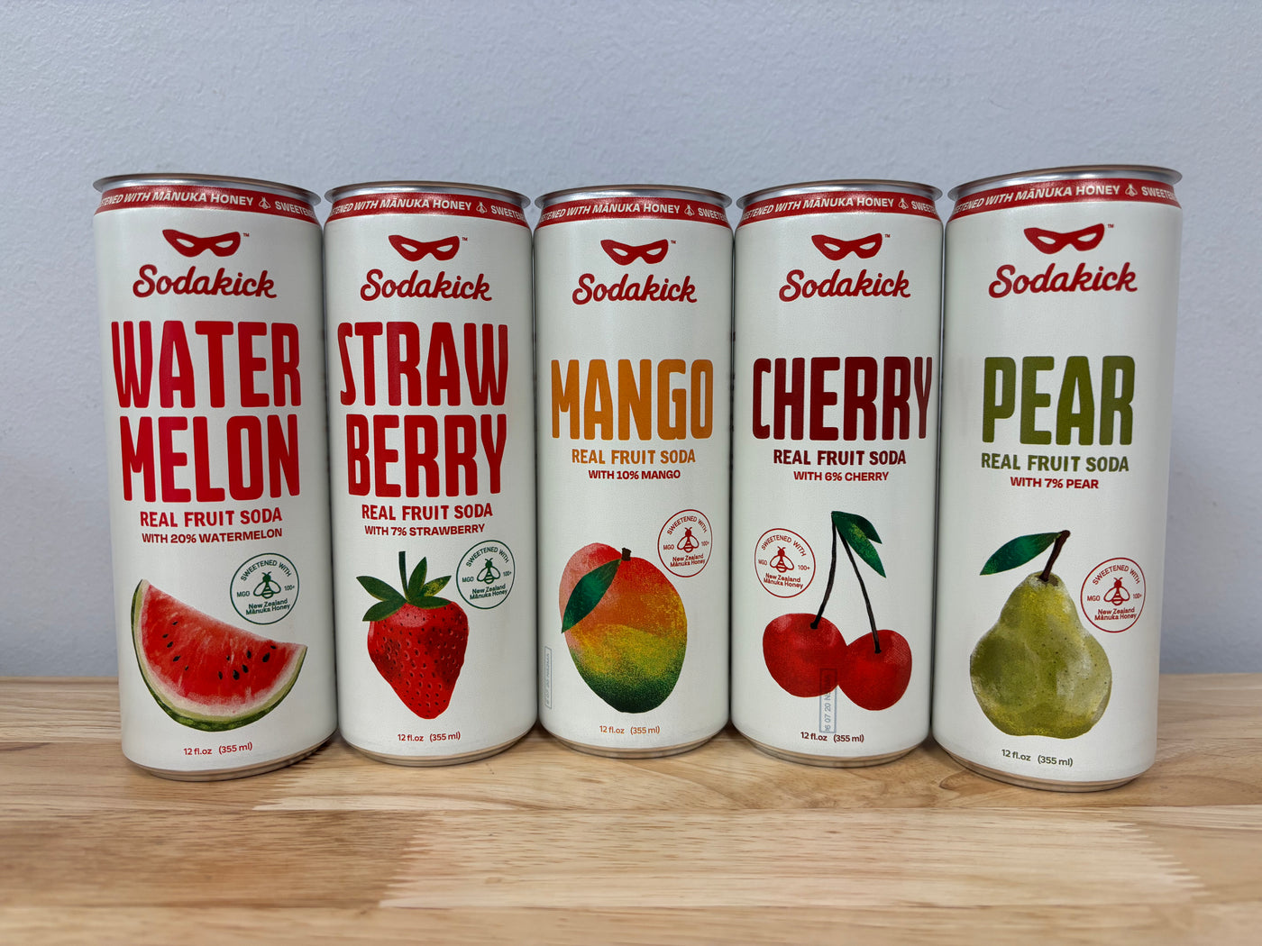 Printed Sodakick sample cans in Watermelon, Strawberry, Mango, Cherry and Pear, lined up on a wooden table