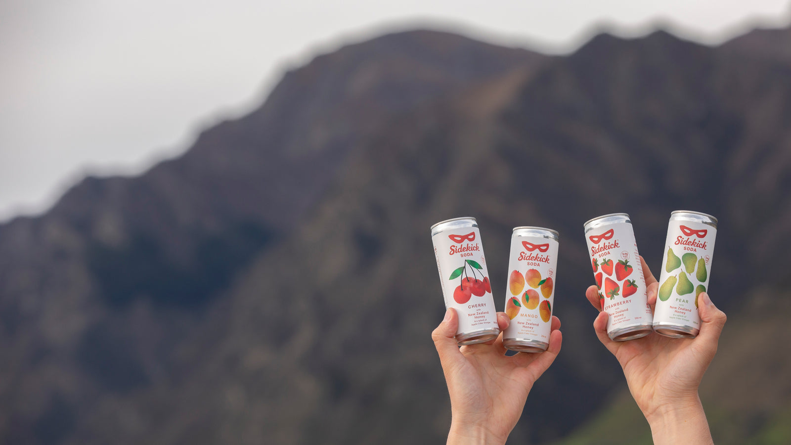 Four Sidekick Soda cans — Cherry, Mango, Strawberry and Pear — held up against a New Zealand mountain backdrop